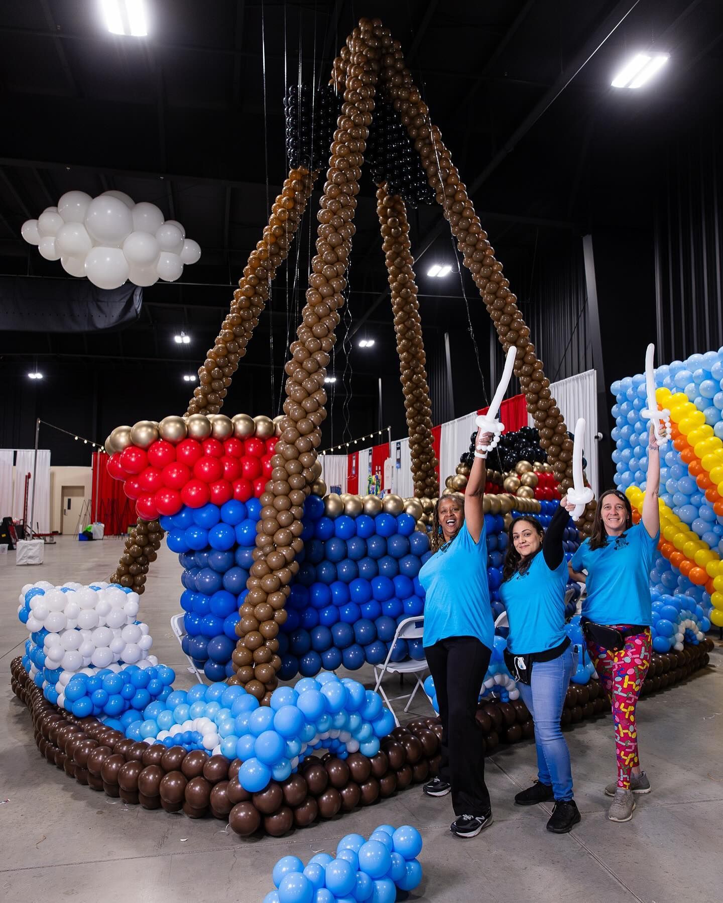 Two people pose beside a giant colorful balloon sculpture in an indoor event hall.