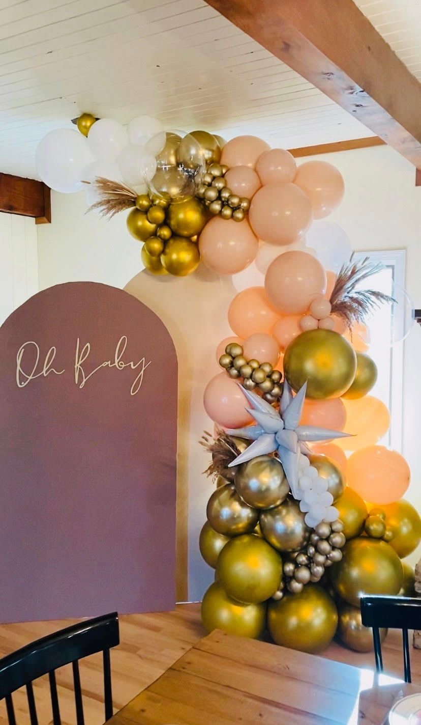 Pastel peach and gold balloon arch beside a maroon “Oh baby” sign at a baby shower or gender reveal