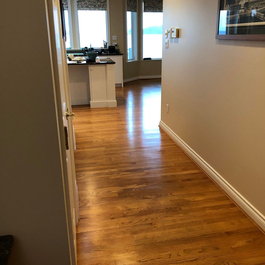 Empty hallway with wood floors leading to a bright kitchen and windows