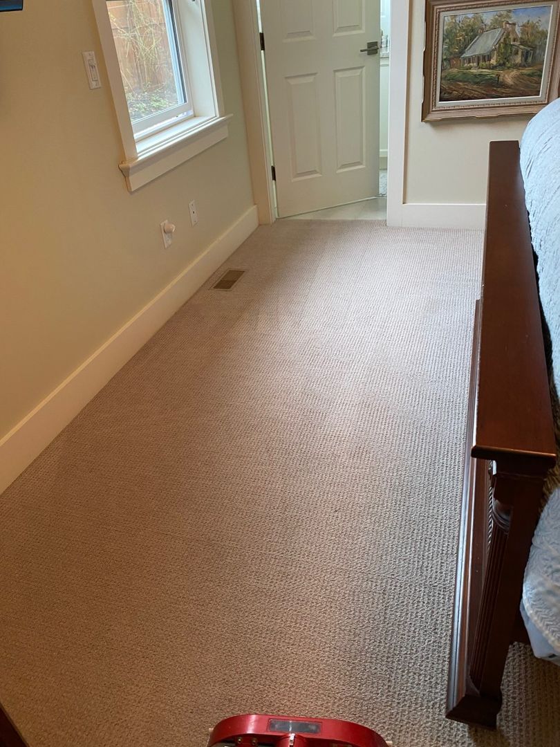 Carpeted hallway with beige walls, a white door, a window, and a wooden bedframe on the right.