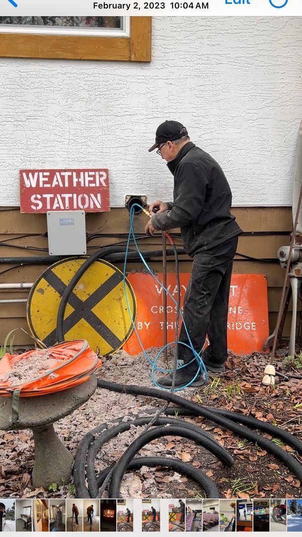 Person pumping water at a weather station with hoses, a yellow reel, and a red sign outside a building