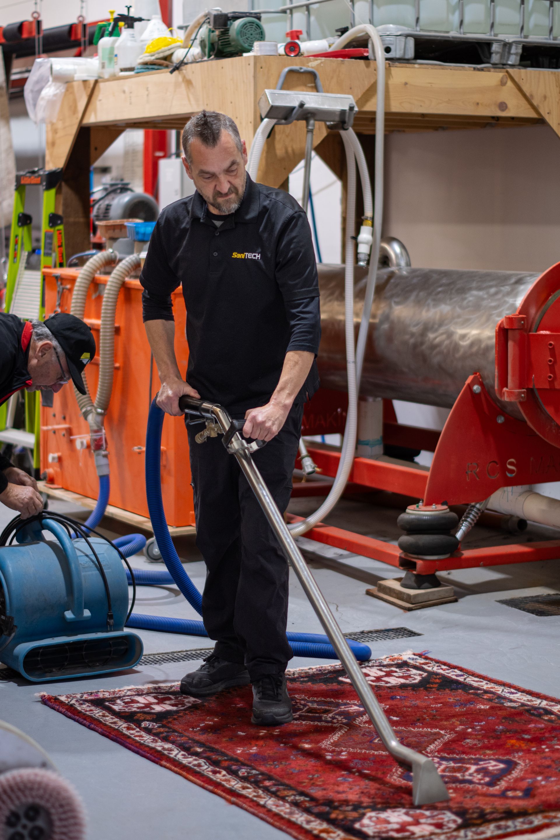 Man cleaning a red rug indoors with a long vacuum wand beside industrial equipment