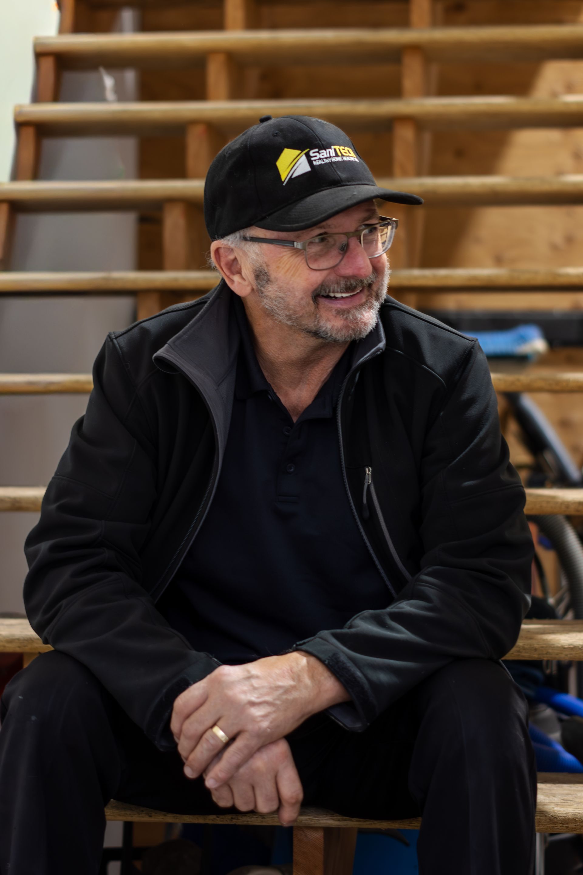 Man in black jacket and cap sitting on wooden bleachers, smiling and looking to the side.