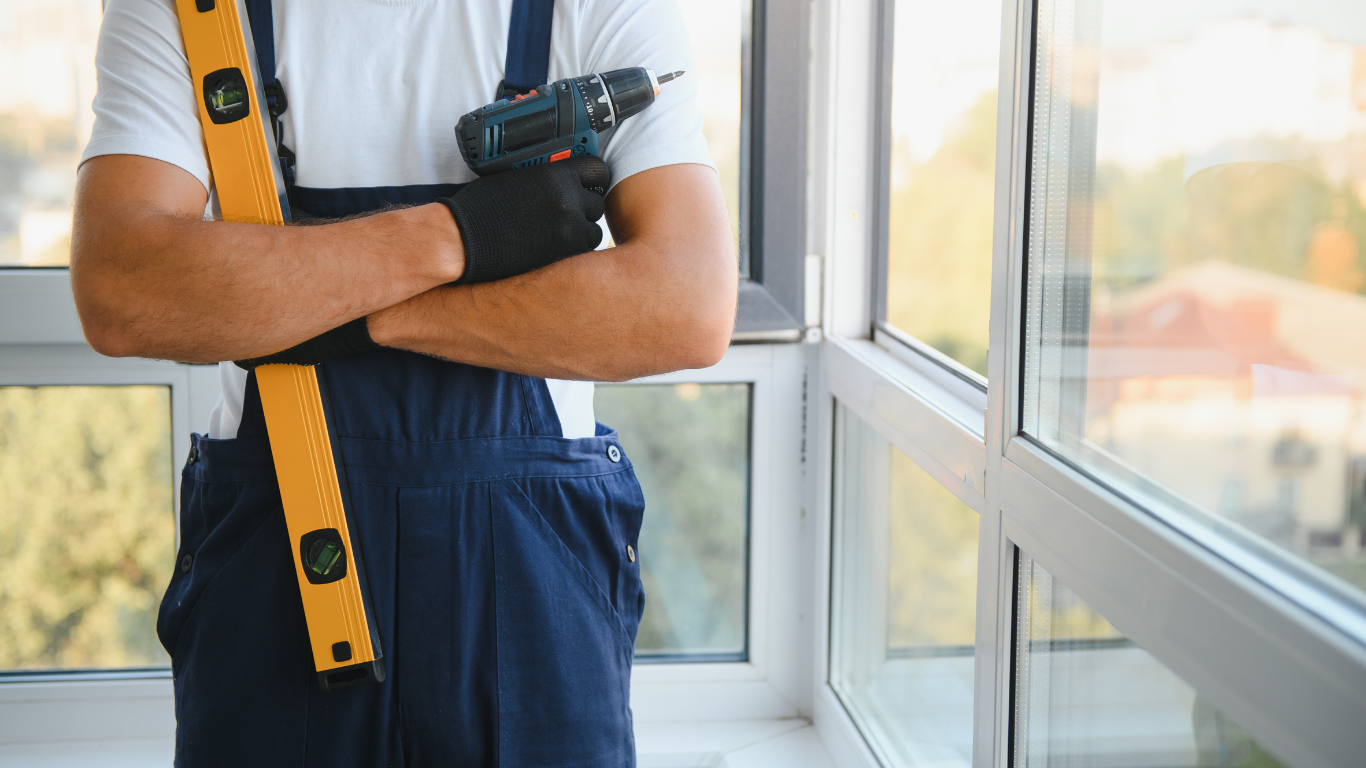 A professional in navy blue overalls holds a yellow spirit level and a cordless drill while standing by a window.
