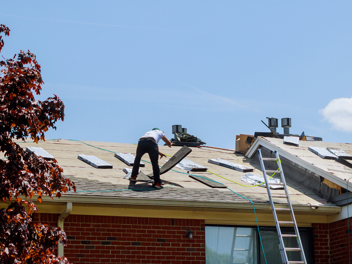 Workers repair the roof of a Victorian-style house under a bright blue sky, with a ladder leaning against the porch.