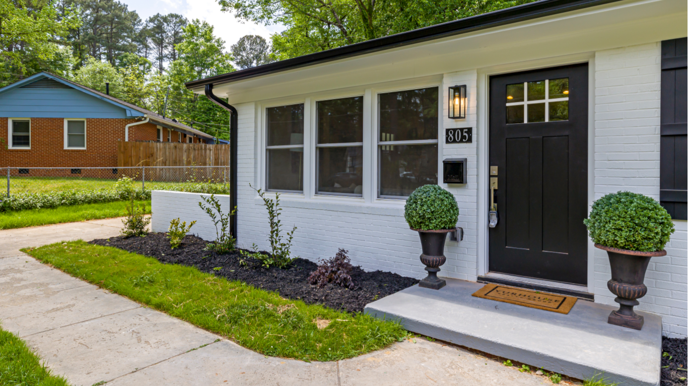 White brick house with black door, windows, and planters. A sidewalk leads to the entrance.