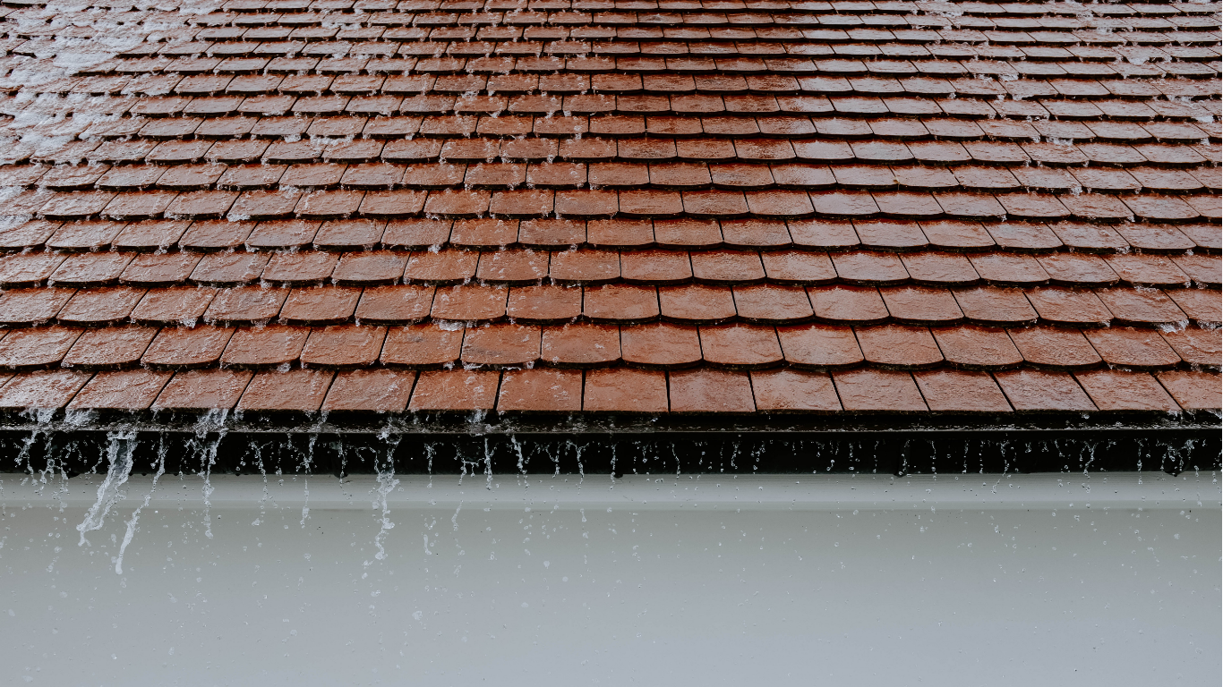 Rain falling off a brown shingled roof into a white gutter.