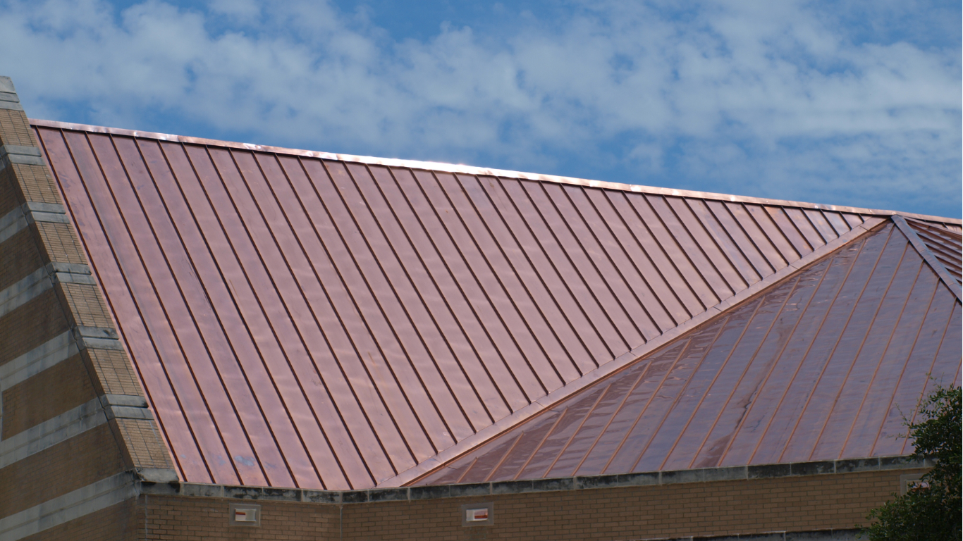Copper-colored metal roof on a brick building, against a blue sky with some clouds.