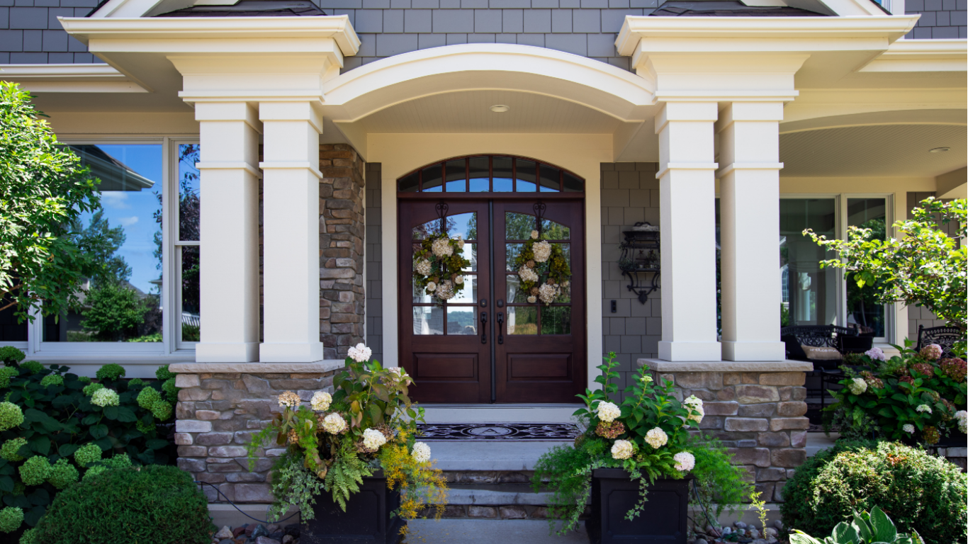 Exterior of a house with a dark wooden door and white pillars framing the entrance. Two flower arrangements sit on either side.