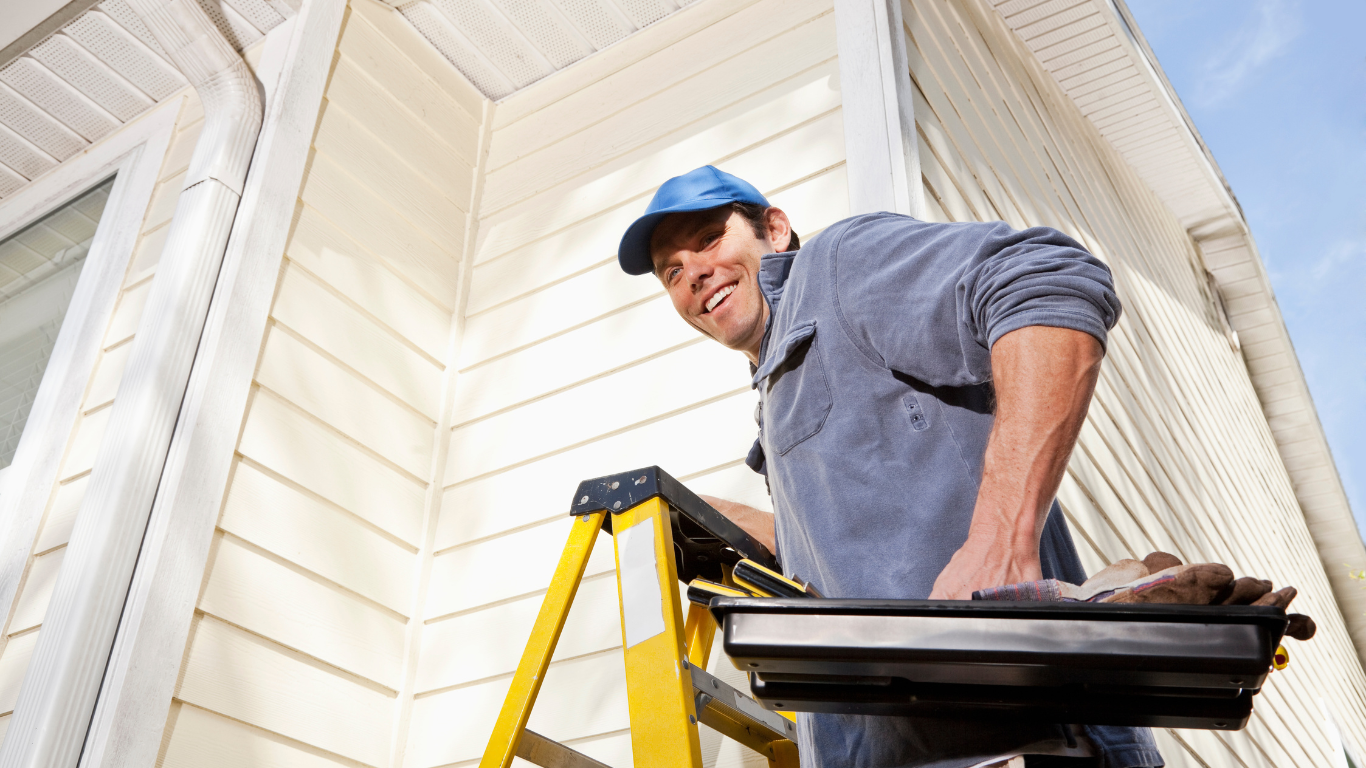 Man on a ladder painting a house exterior, smiling. He wears a blue hat and holds a paint tray.
