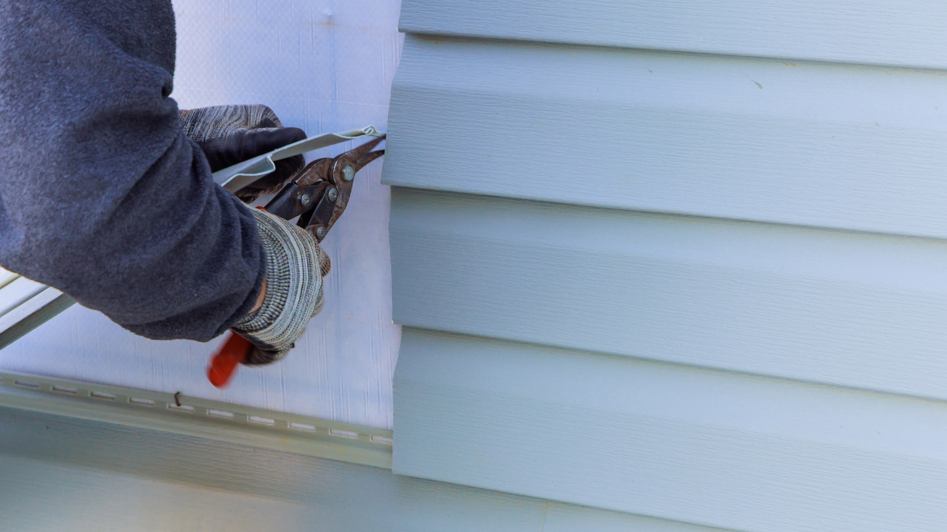 Person in gloves cutting vinyl siding with shears near a house.