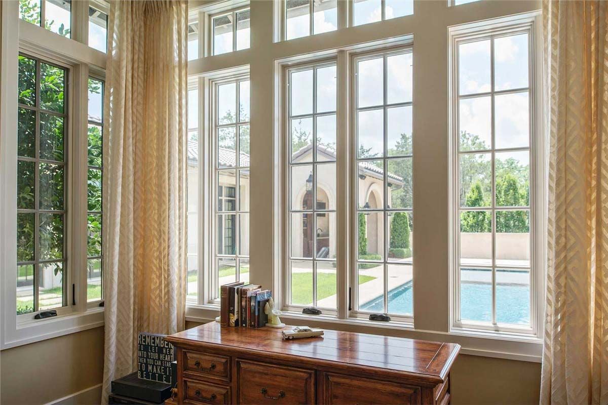 Sunlit room with large windows overlooking a backyard pool and a decorative wooden desk. Cream-colored curtains.
