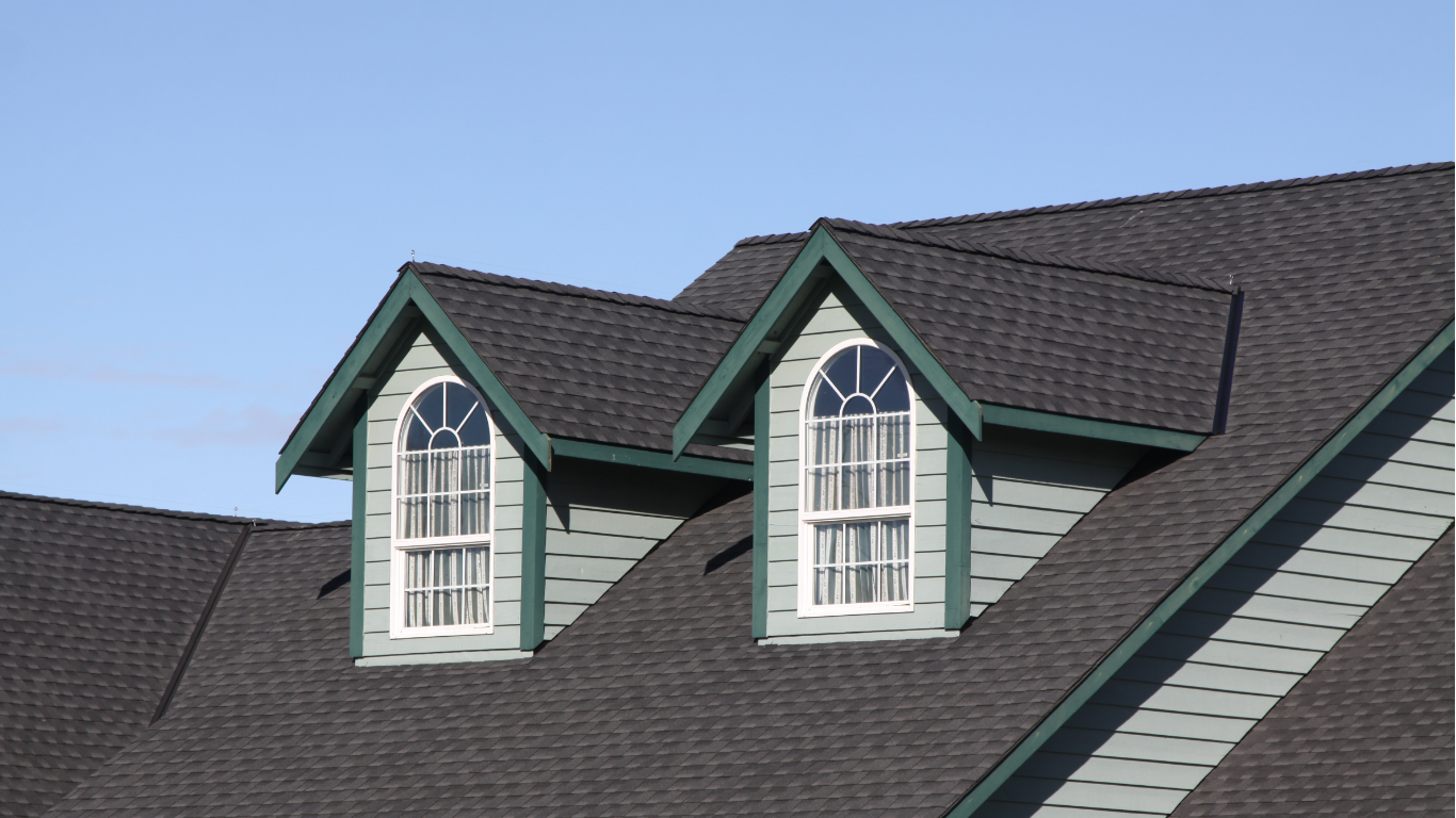 Workers repair the roof of a Victorian-style house under a bright blue sky, with a ladder leaning against the porch.