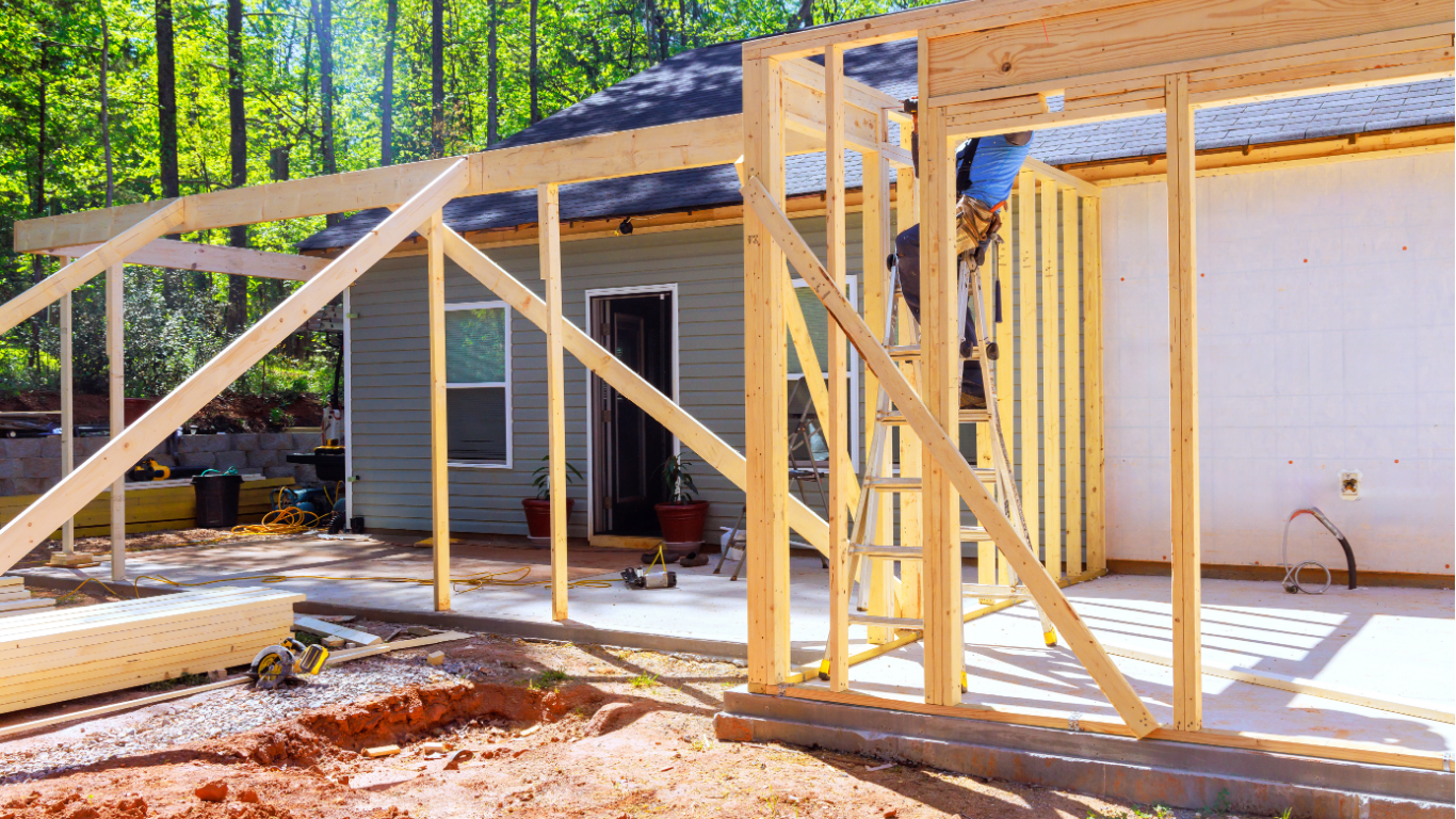 Workers repair the roof of a Victorian-style house under a bright blue sky, with a ladder leaning against the porch.