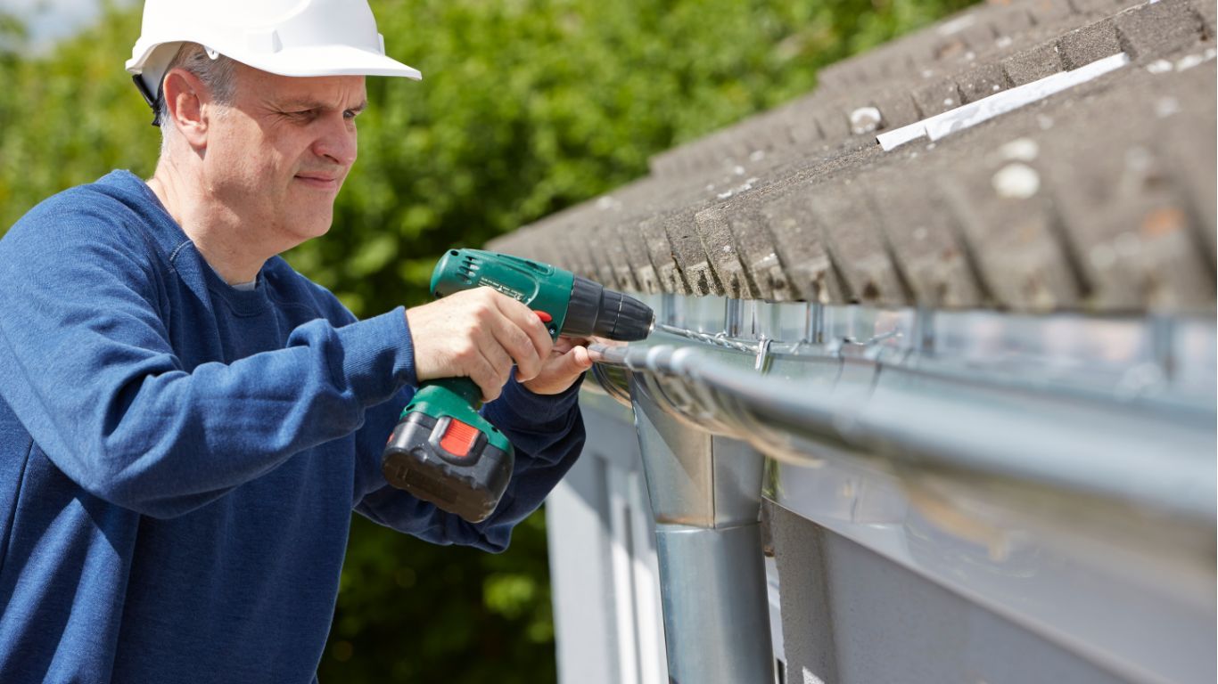 A person in a white hard hat uses a green cordless drill to install a metal gutter on a building exterior.