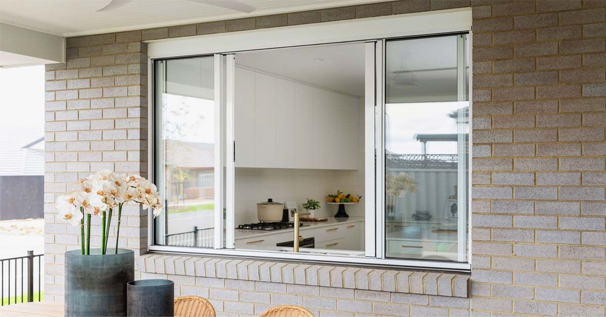 Large window in a brick wall. View into a kitchen. Table with flowers in foreground.