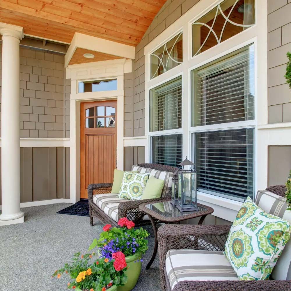 A covered porch with wicker furniture and flower pots. Tan siding, wooden door, and large windows are visible.