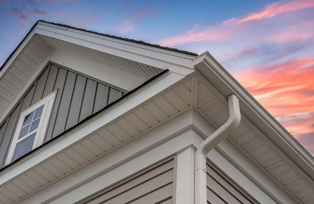 Exterior view of a house with white trim, gray siding, and a gutter, against a colorful sunset.