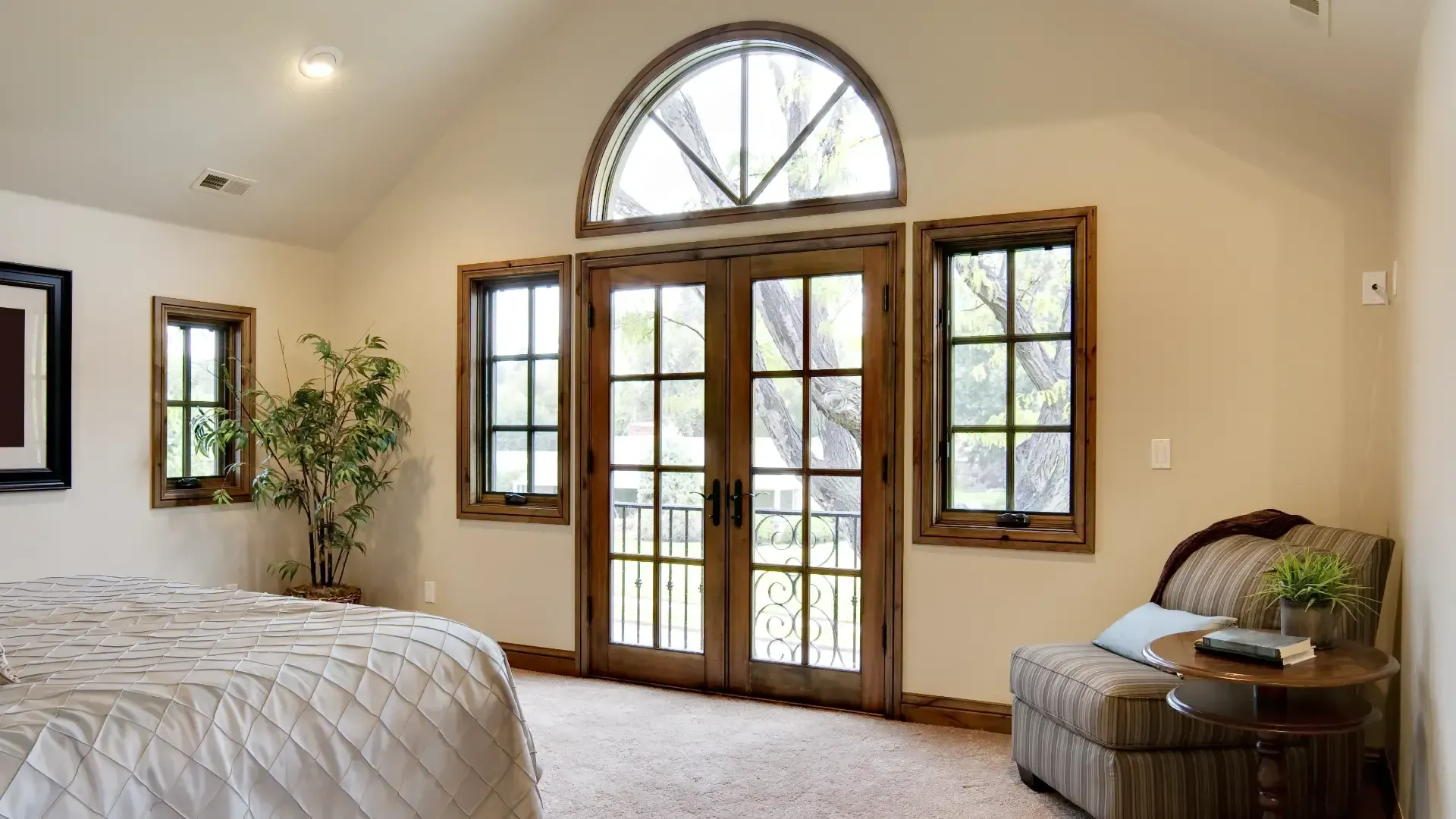 Bedroom with light-colored walls, large arched window, French doors, and a chair near a small table.