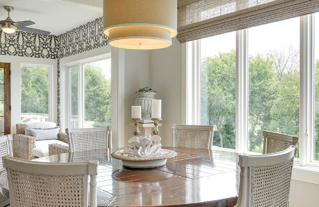 A round wooden dining table surrounded by light-colored wicker chairs in a bright sunroom with large windows.