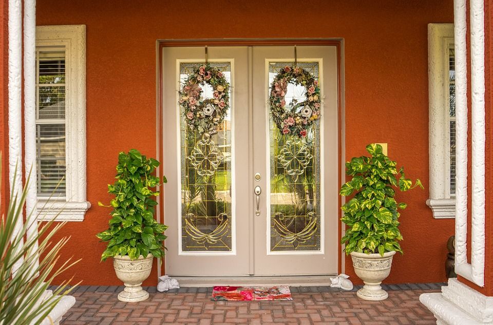 Double glass doors with wreaths, flanked by potted plants, and windows on a reddish-orange house.