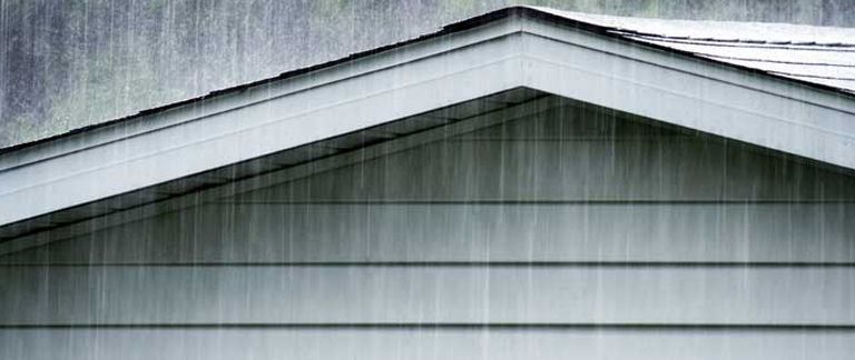Rain falls against the gray siding and overhanging roof of a house.