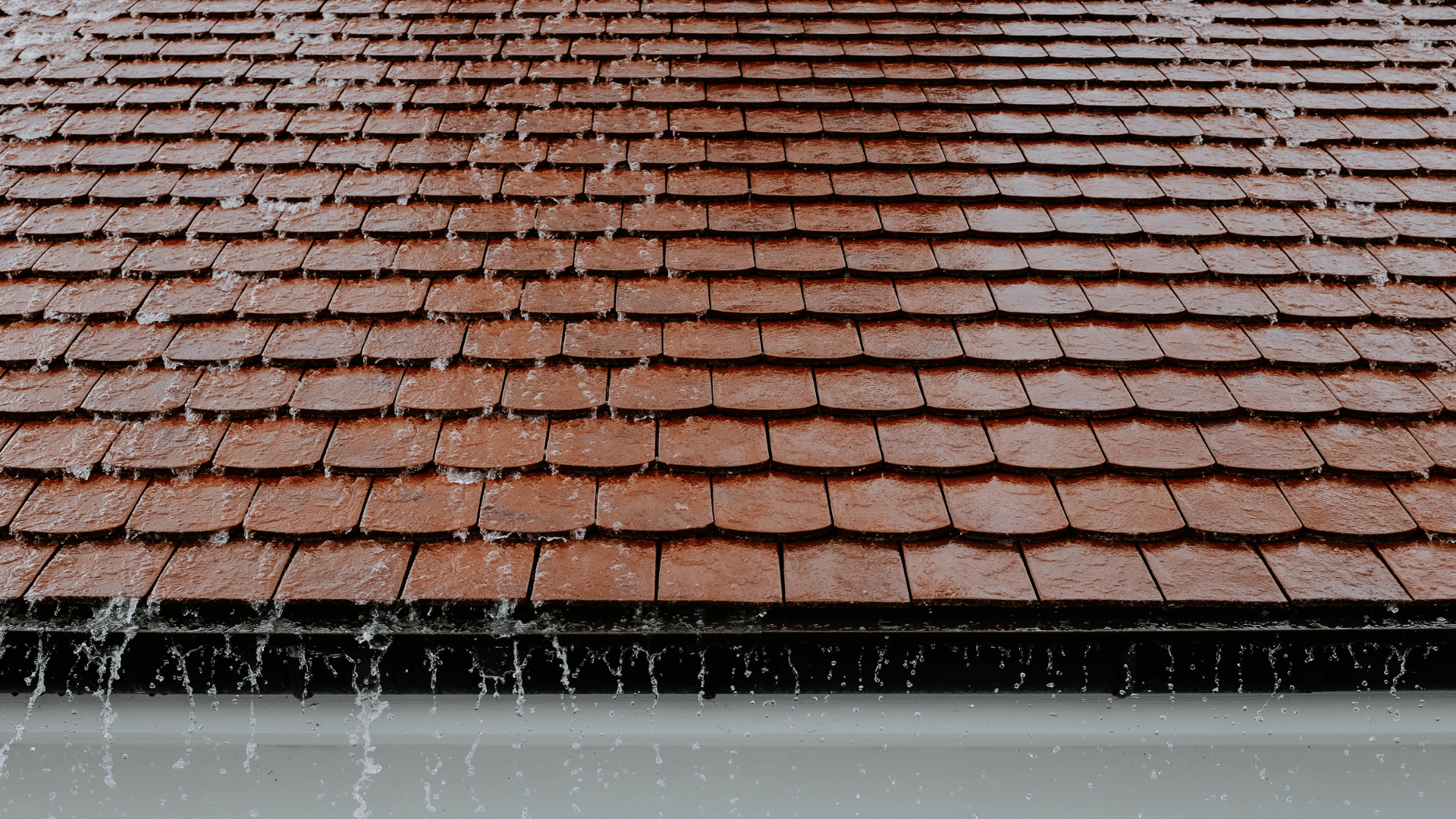 Rain falling on a brown tiled roof with a white gutter.