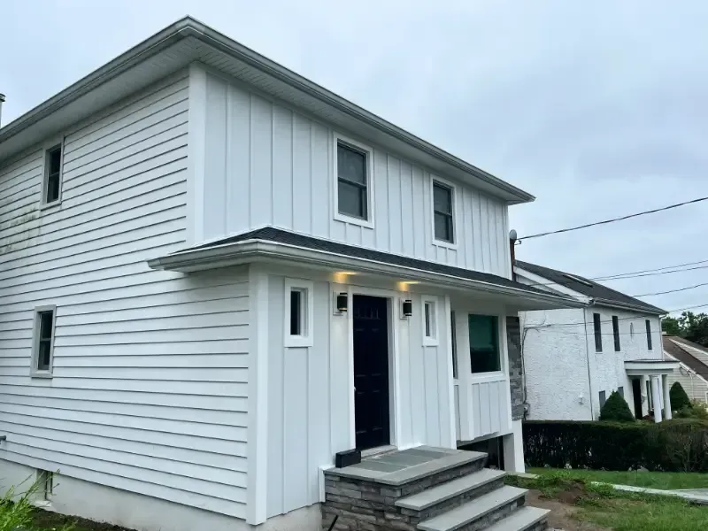 White two-story house with vertical and horizontal siding, stone steps leading to a dark door under a small overhang.