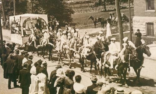 A historical photograph of a street parade with horse-drawn carriages and riders in white uniforms, viewed by a crowd.