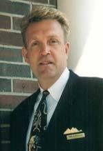 A person in a dark pinstriped suit, patterned tie, and name tag standing against a dark brick background.