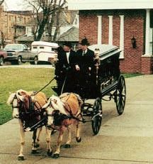 Two people in formal suits sit on a horse-drawn hearse, being pulled by two horses on a paved driveway near a brick building.