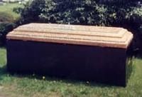 A rectangular, above-ground grave marker with a dark base and a layered, light-colored stone top, situated on a lawn.