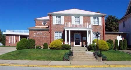 A two-story brick funeral home with white trim, a columned porch, manicured shrubs, and a lawn under a clear blue sky.