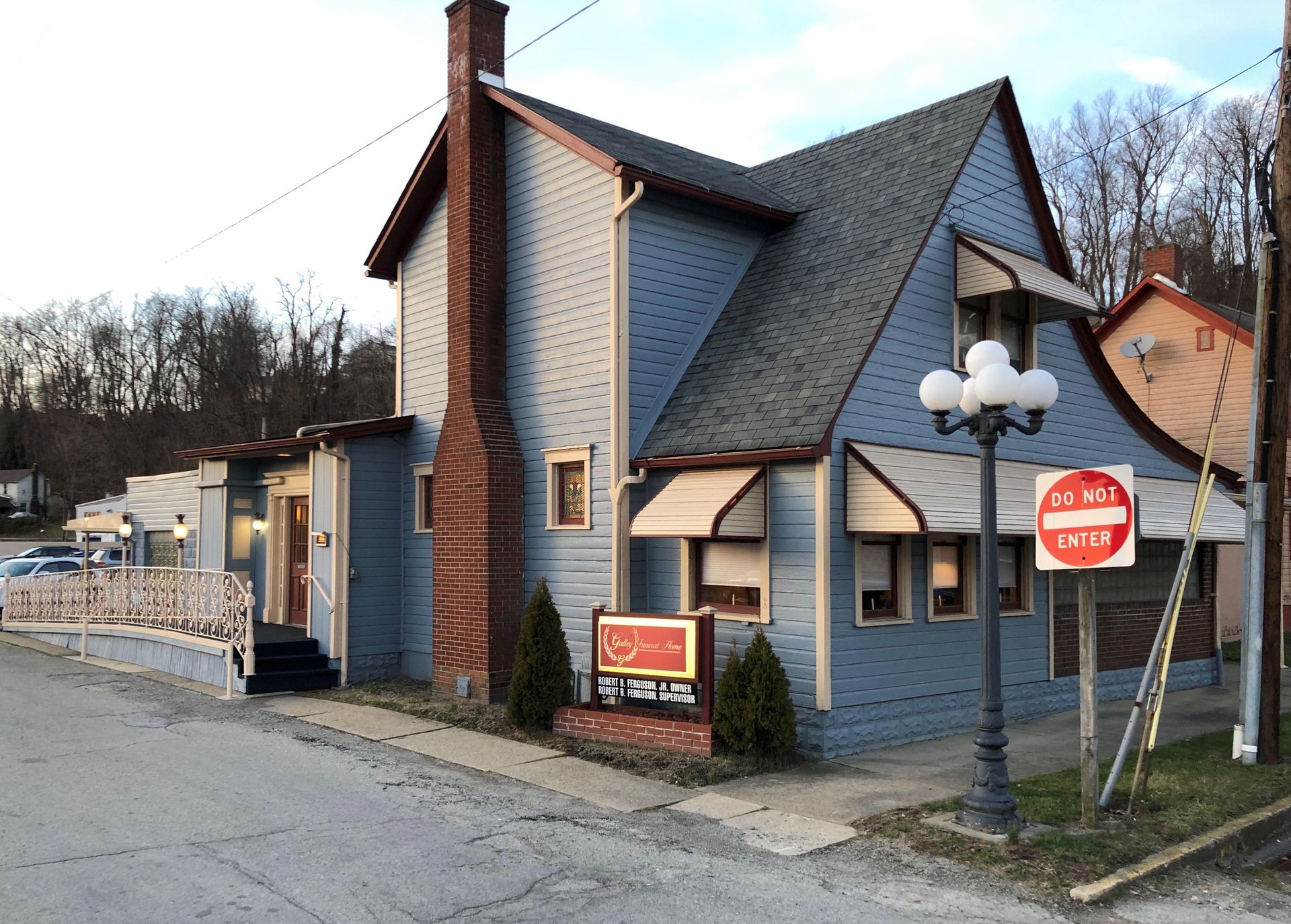 A blue, two-story house with a brick chimney and metal awnings sits beside a street with a 