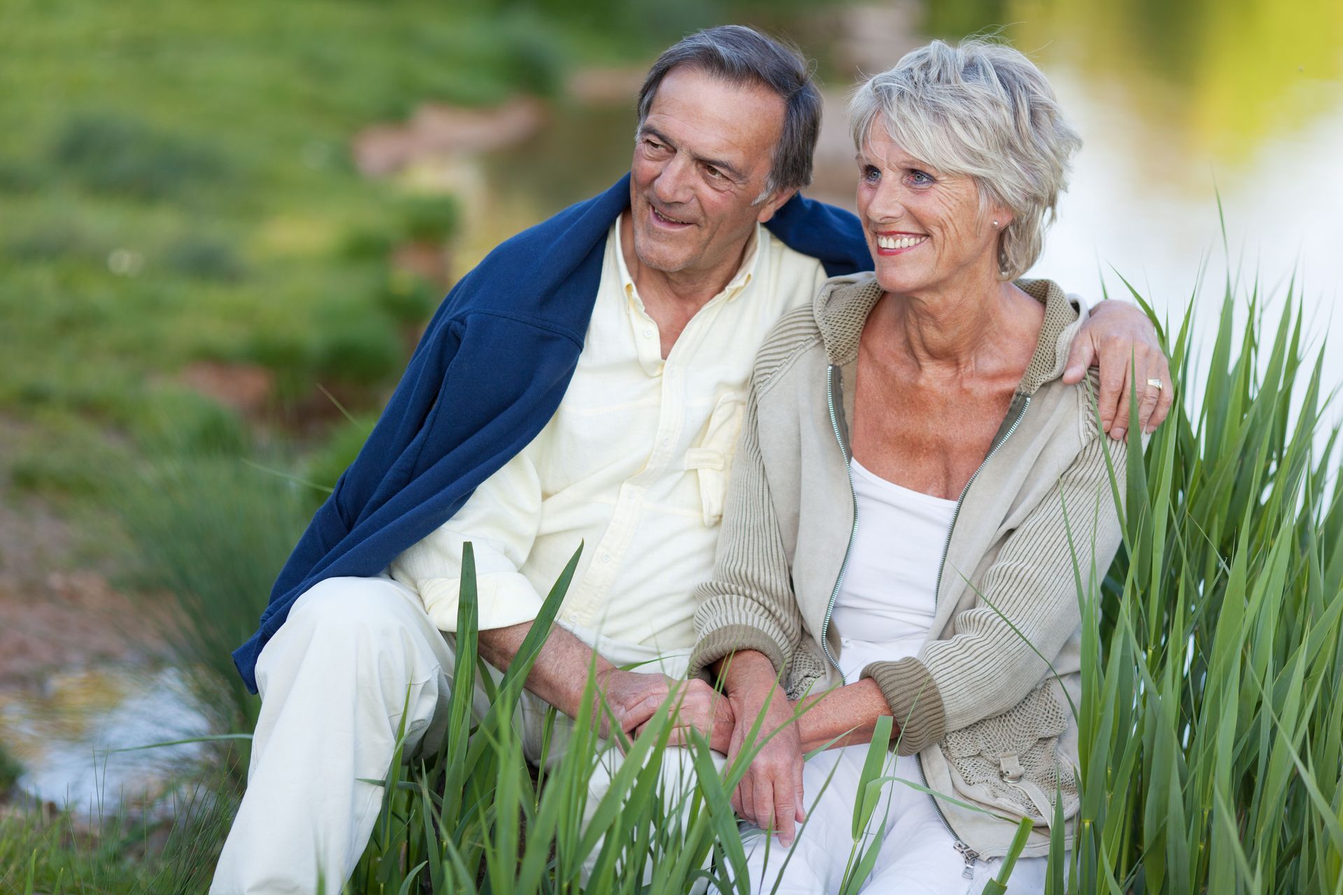 A smiling couple sits together in tall grass by a body of water, wearing casual light-colored clothing.