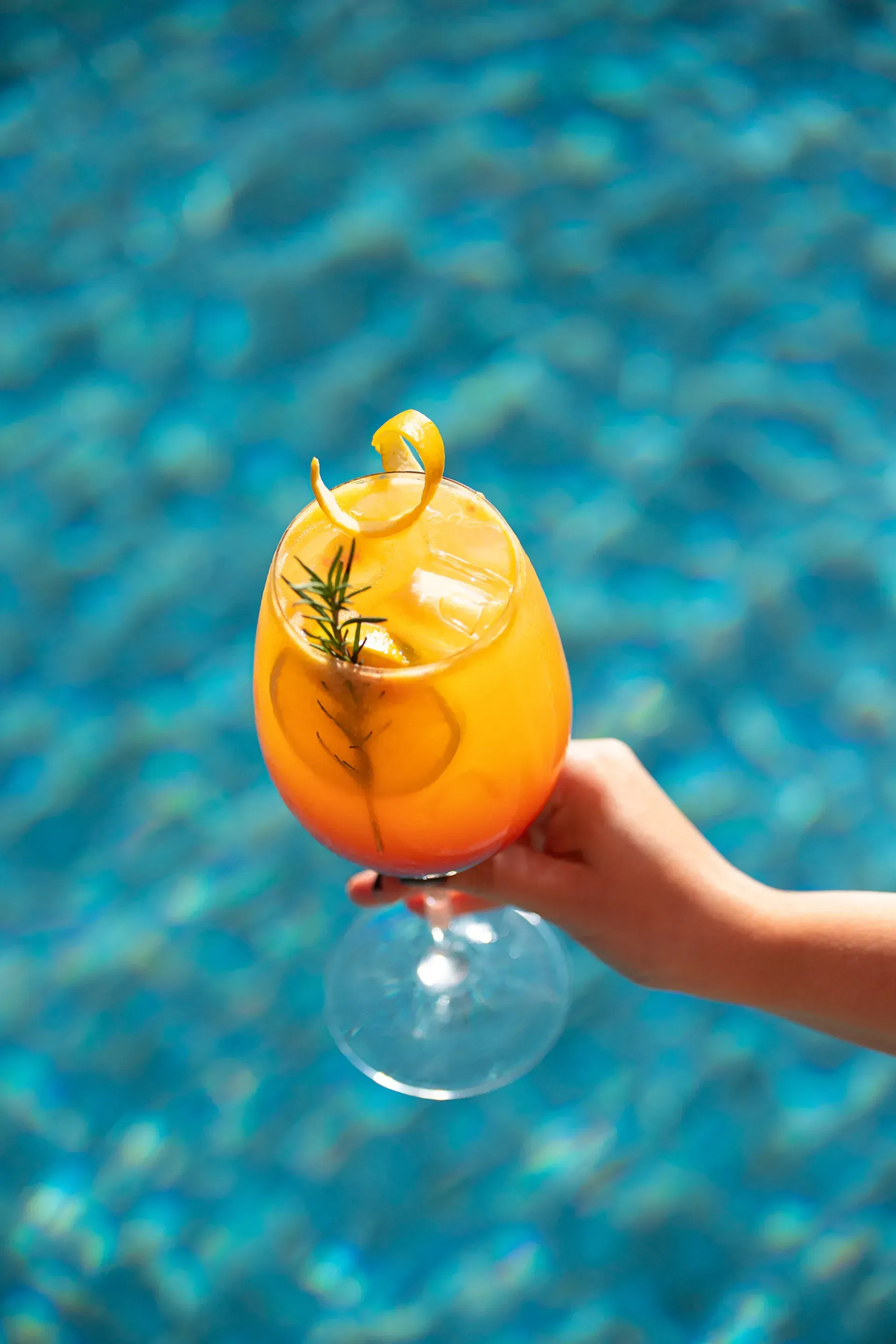 Mão segurando um coquetel de laranja com ervas à beira de uma piscina azul.