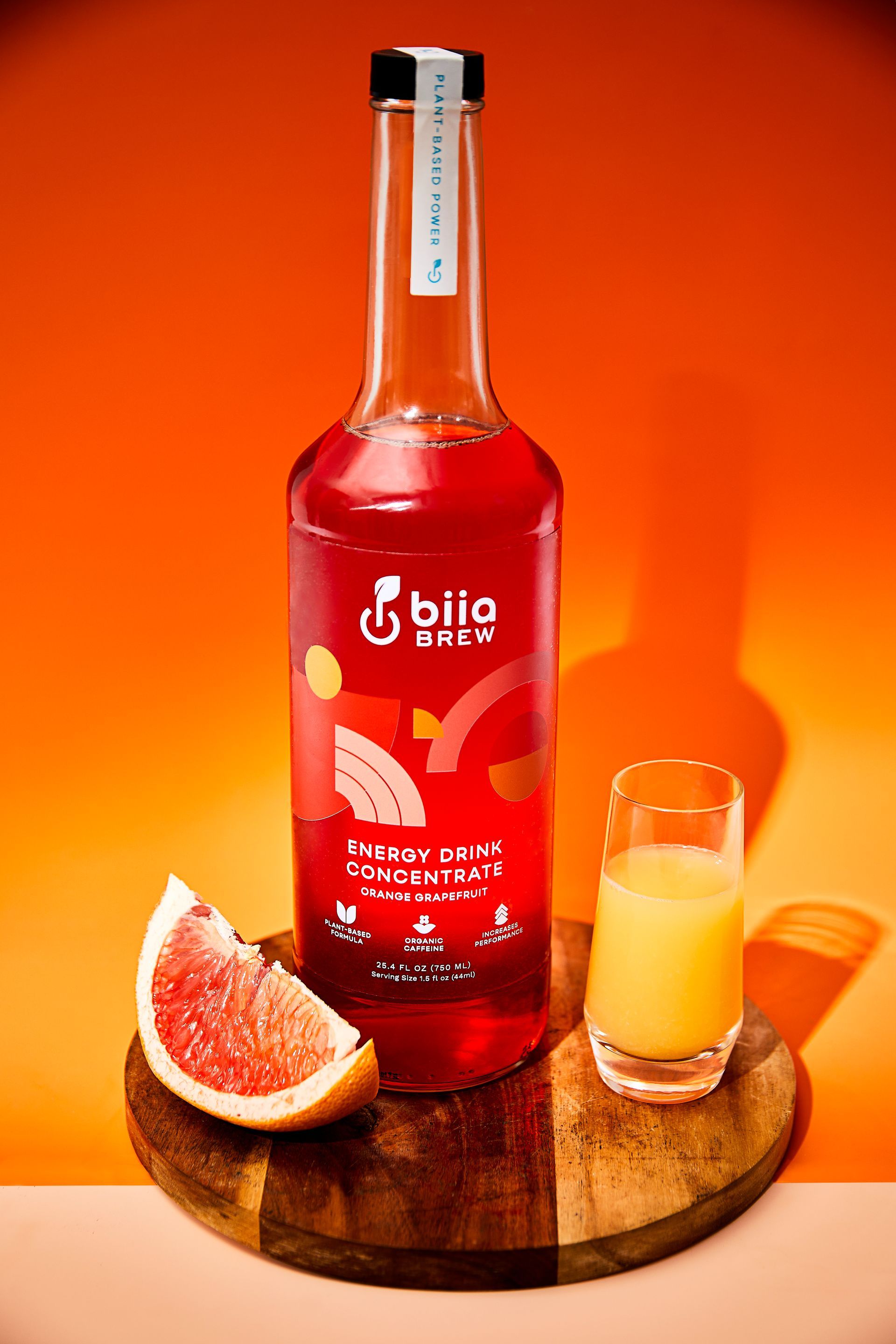 Red beverage bottle with grapefruit and a small filled glass on a wooden board against an orange background