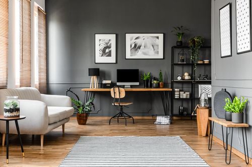 Modern home office with wooden desk, chair, plants, gray walls, and striped rug in a bright living space