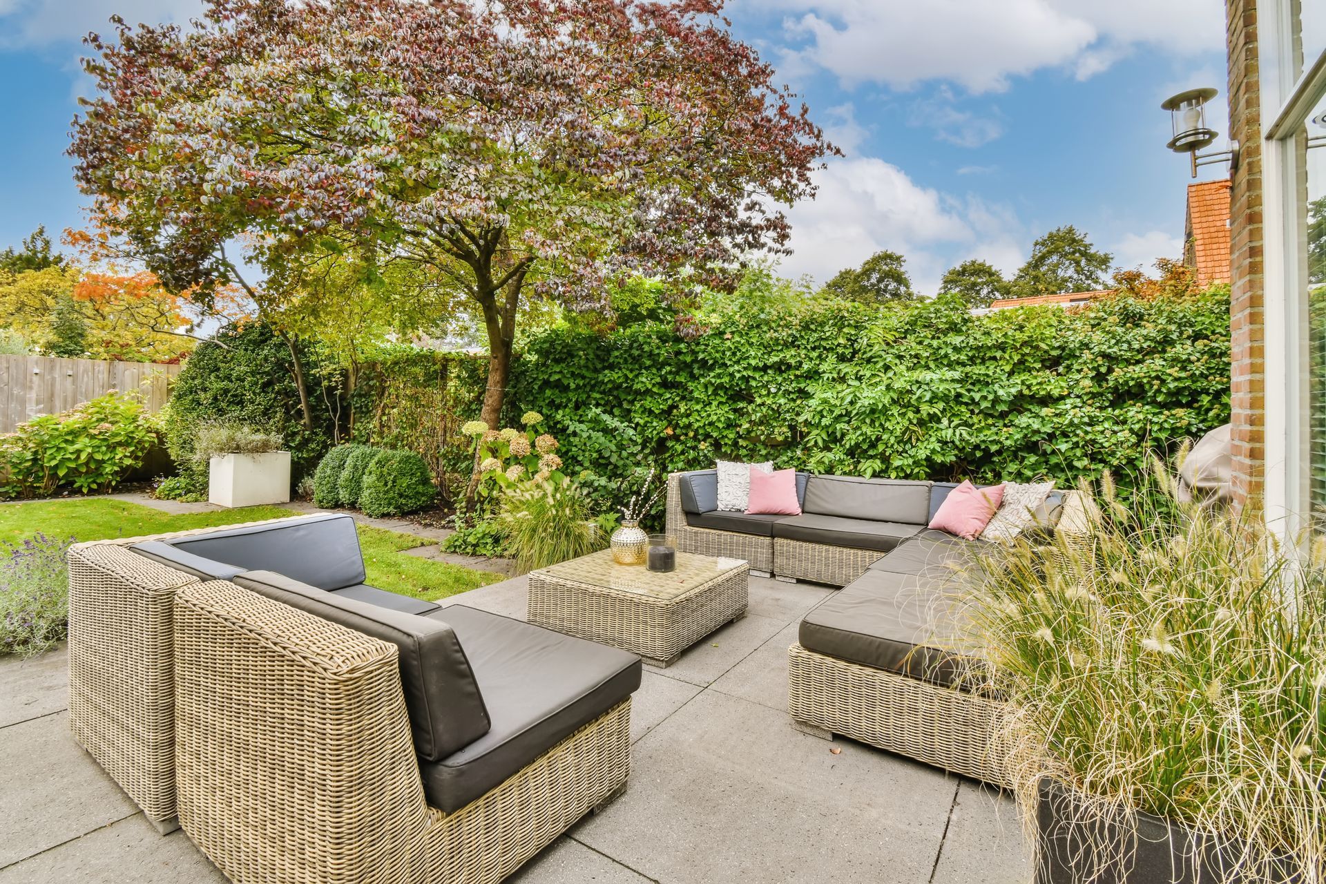 Outdoor patio with wicker seating, a coffee table, and green hedges under a tree