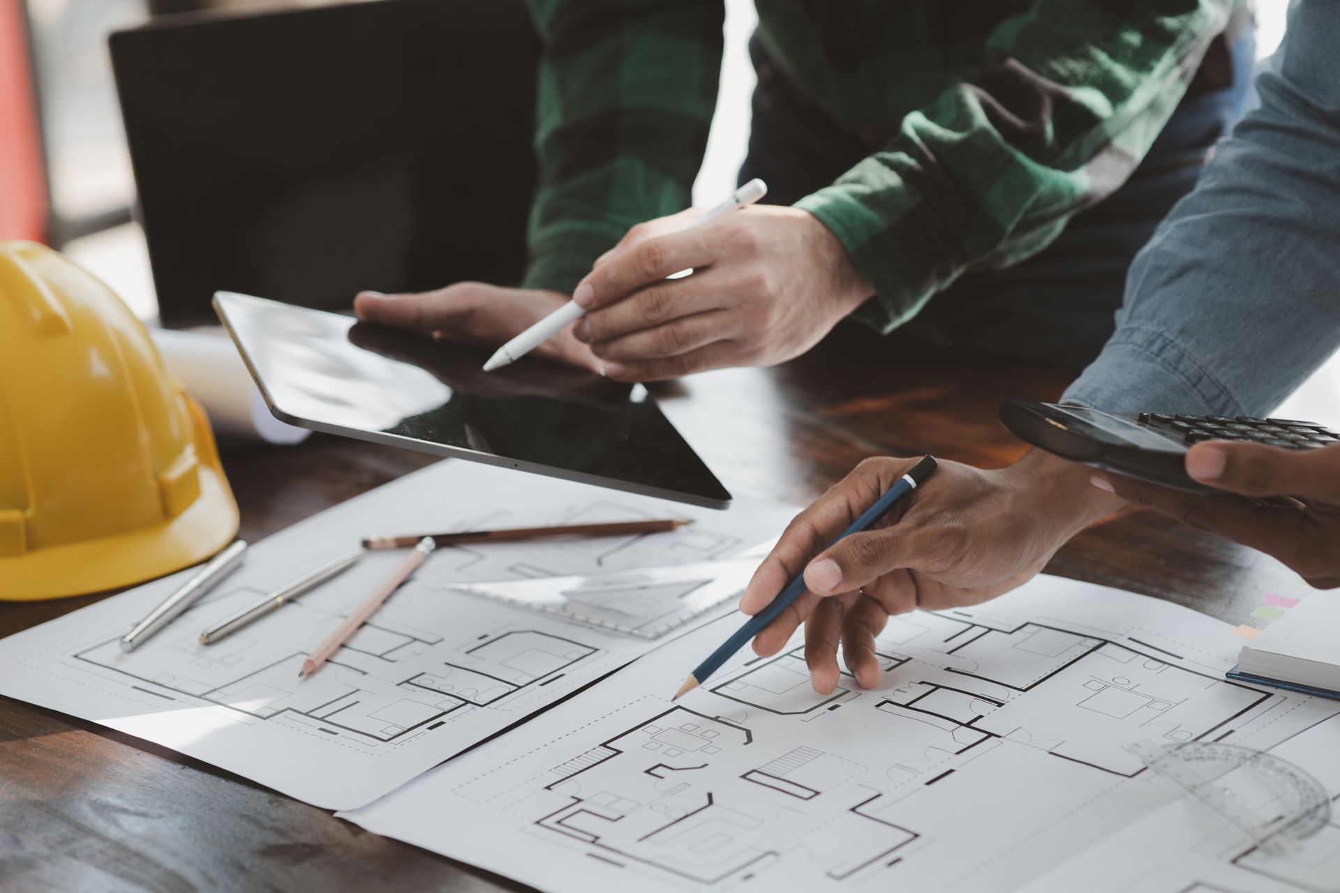 Hands reviewing building plans on a table with a yellow hard hat and pencil sketches
