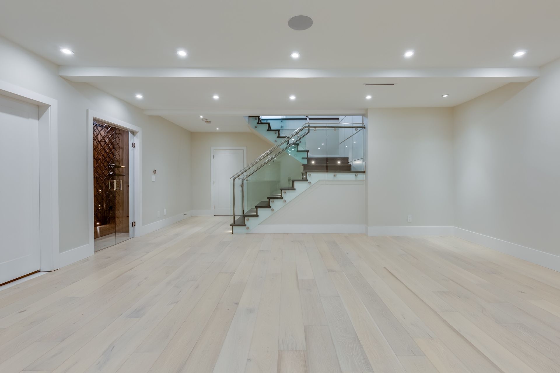 Empty modern living room with light wood floors, recessed lights, and a glass staircase.