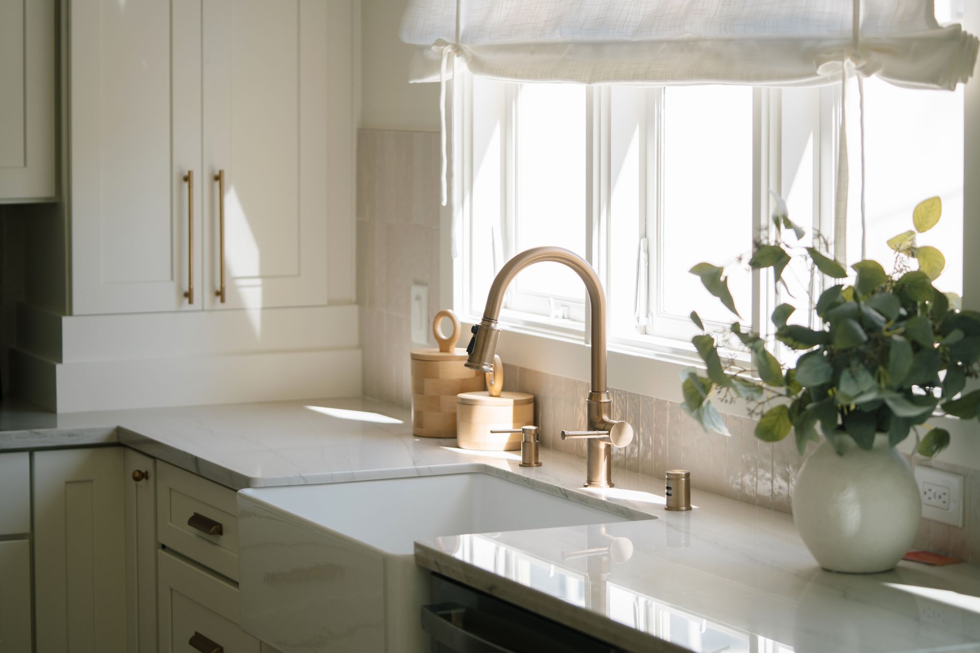 Bright kitchen sink by a sunlit window, with white cabinets and a vase of greenery.