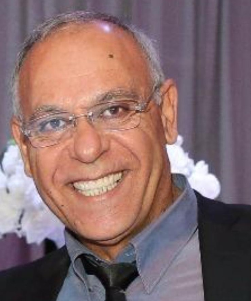 גבר עם משקפיים מחייך, לובש ז'קט חליפה שחור וחולצה עם צווארון פתוח.