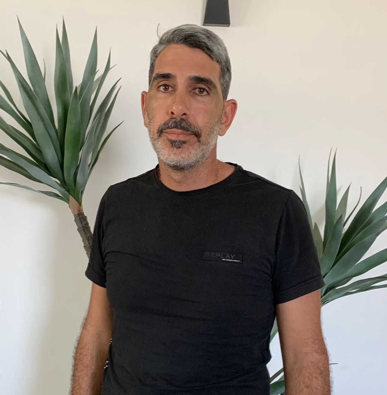 גבר עם שיער וזקן מאפירים בחולצה שחורה, עומד מול צמח ירוק.