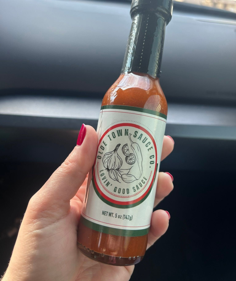 Hand holding a hot sauce bottle with a white label and dark green cap in a car interior