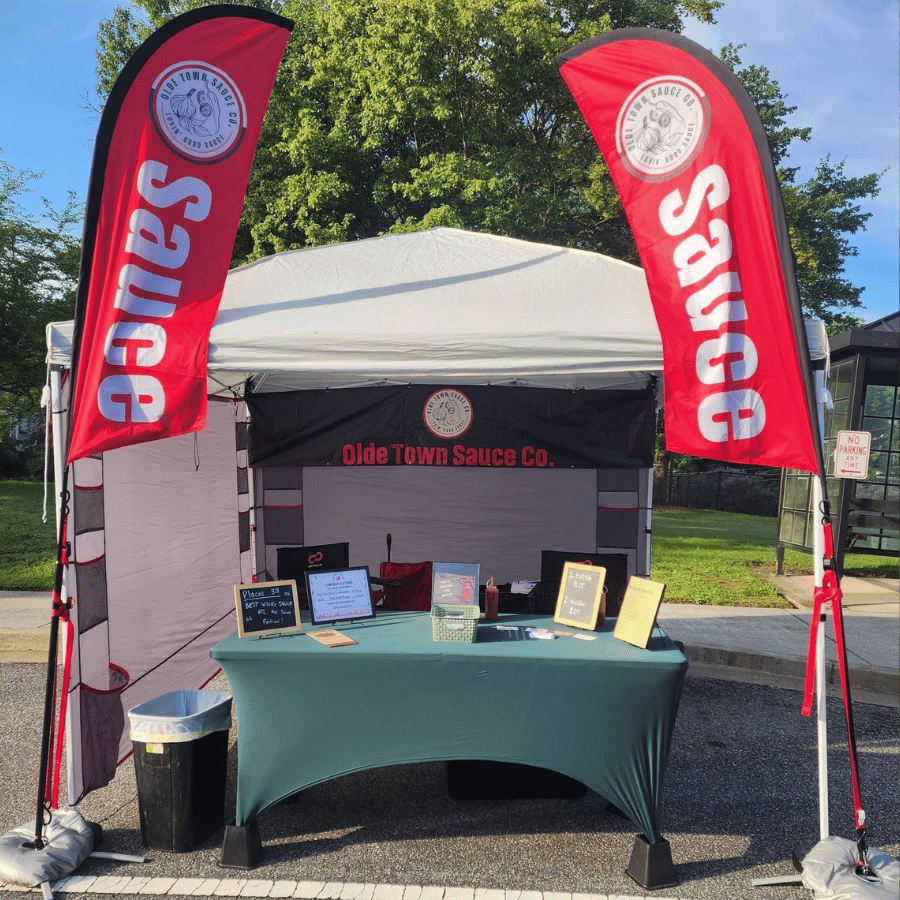 Outdoor vendor booth with red “Sauce” flags and a display table under a white canopy.