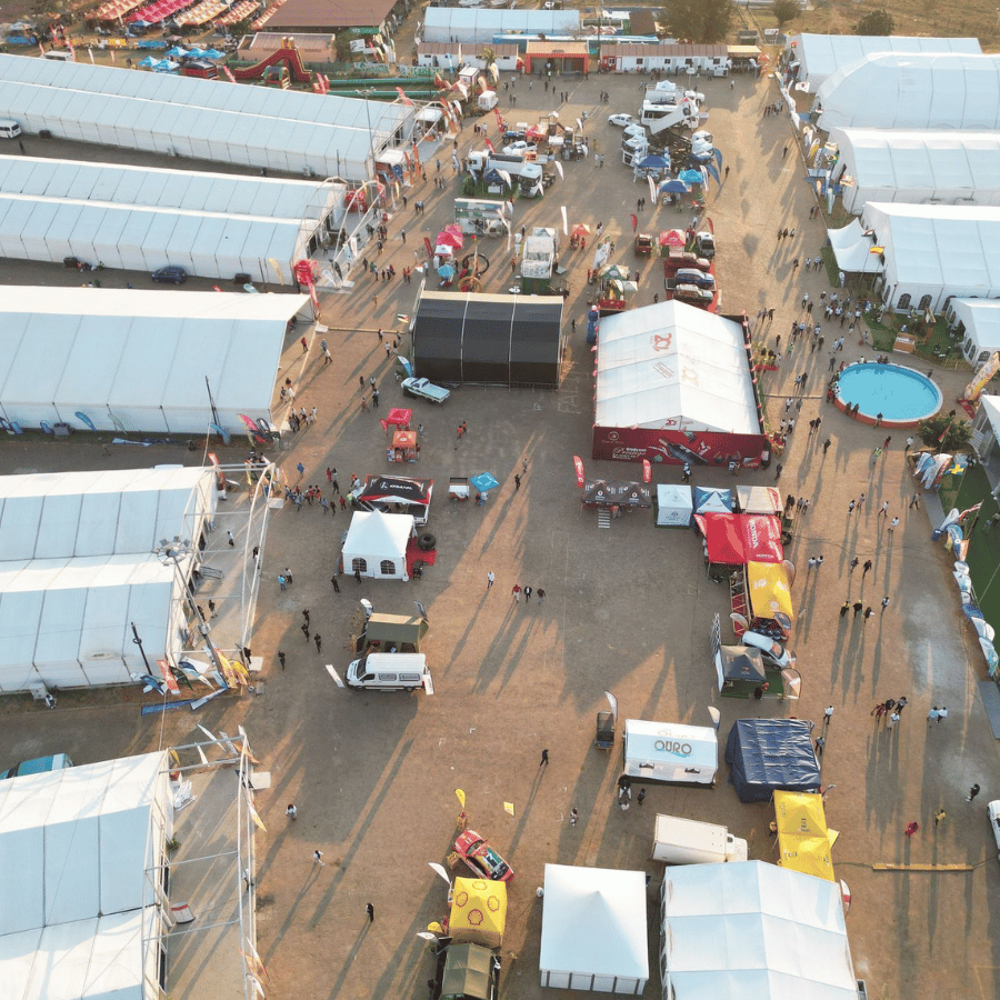 Aerial view of a crowded outdoor camp with white tents, vehicles, and dirt roads.