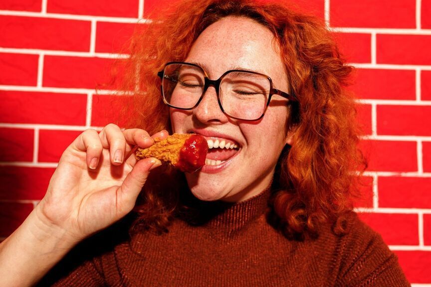Person with glasses eating a chicken wing against a red brick wall.