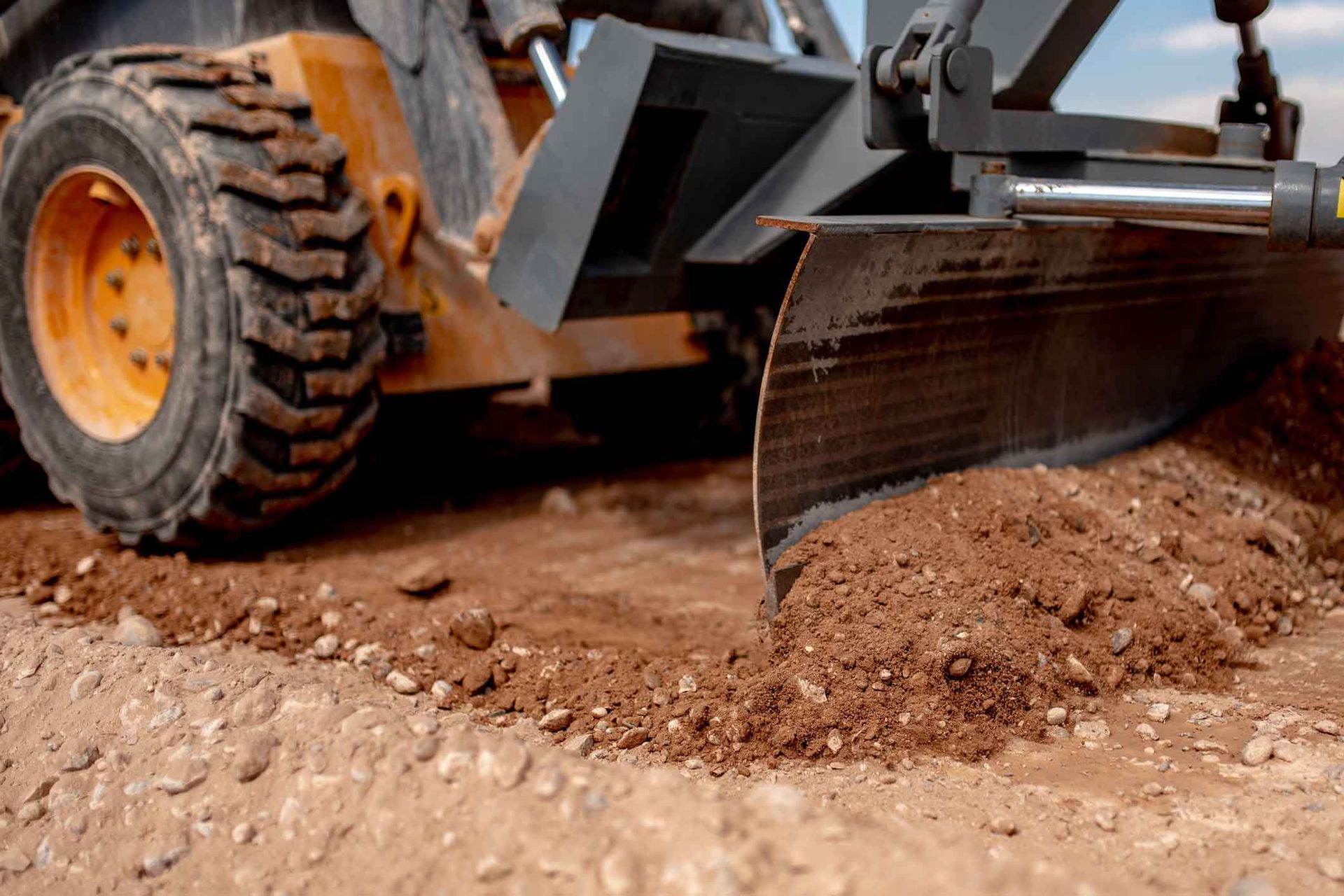 A motor grader blade scraping dirt on a construction site with a wheel in the foreground.