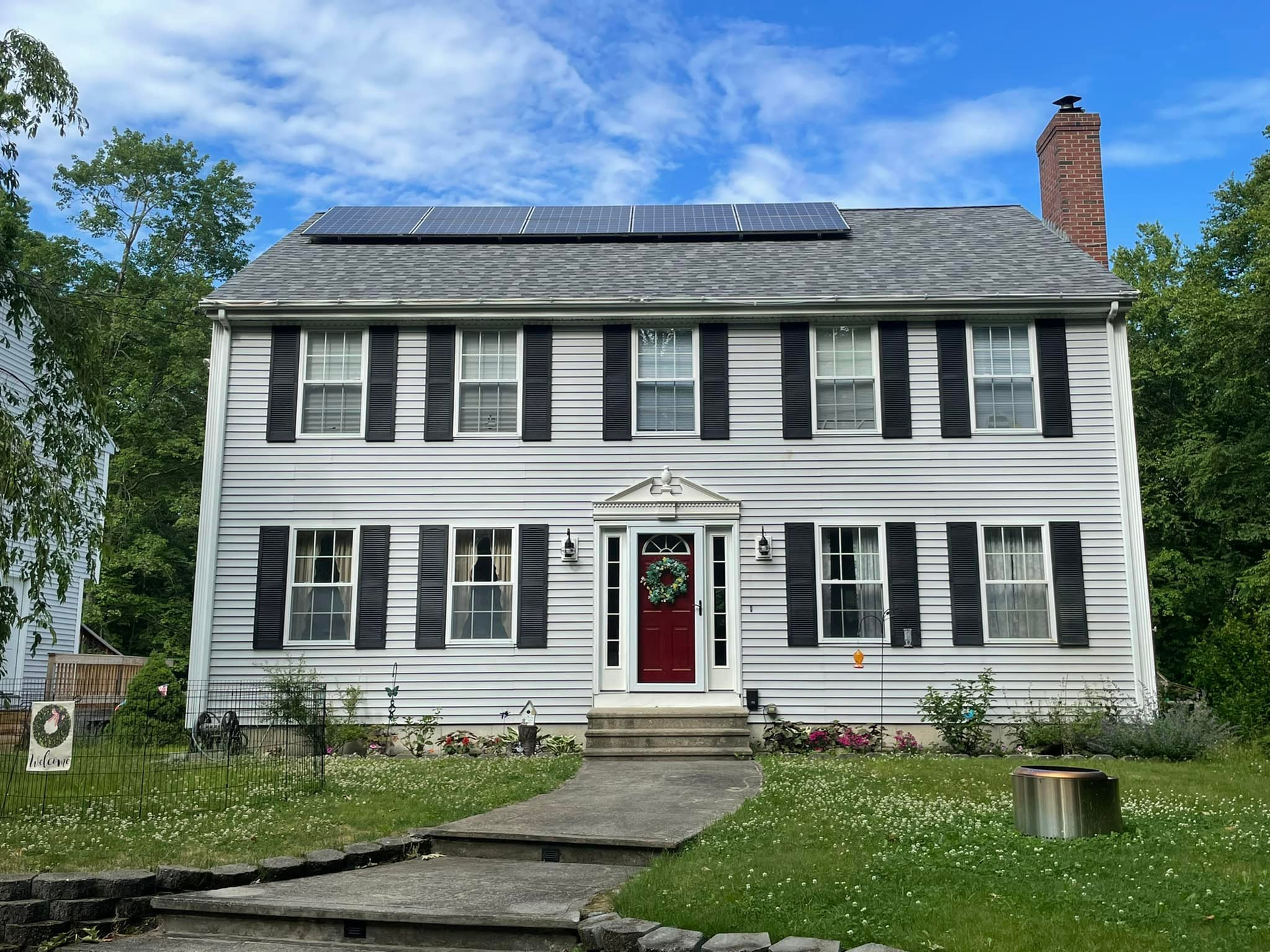 White colonial-style house with black shutters, red front door, and solar panels on the roof under a blue sky